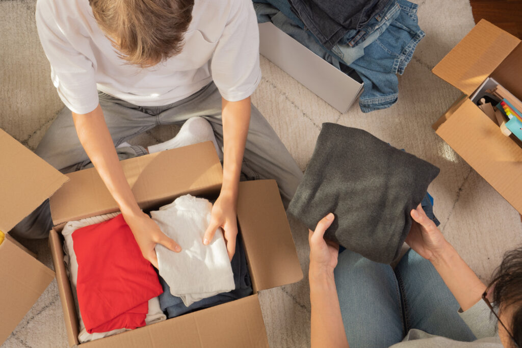 Woman and child sorting clothes and packing into cardboard box. Donations for charity, help low income families, declutter home, sell online, moving moving into new home, recycling, sustainable living.